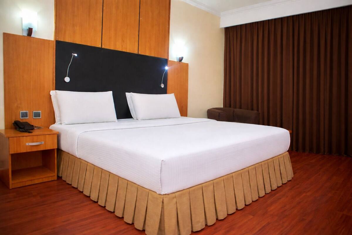 Quarto de hotel com uma cama grande, cabeceira de madeira, mesas de cabeceira e cortina marrom.