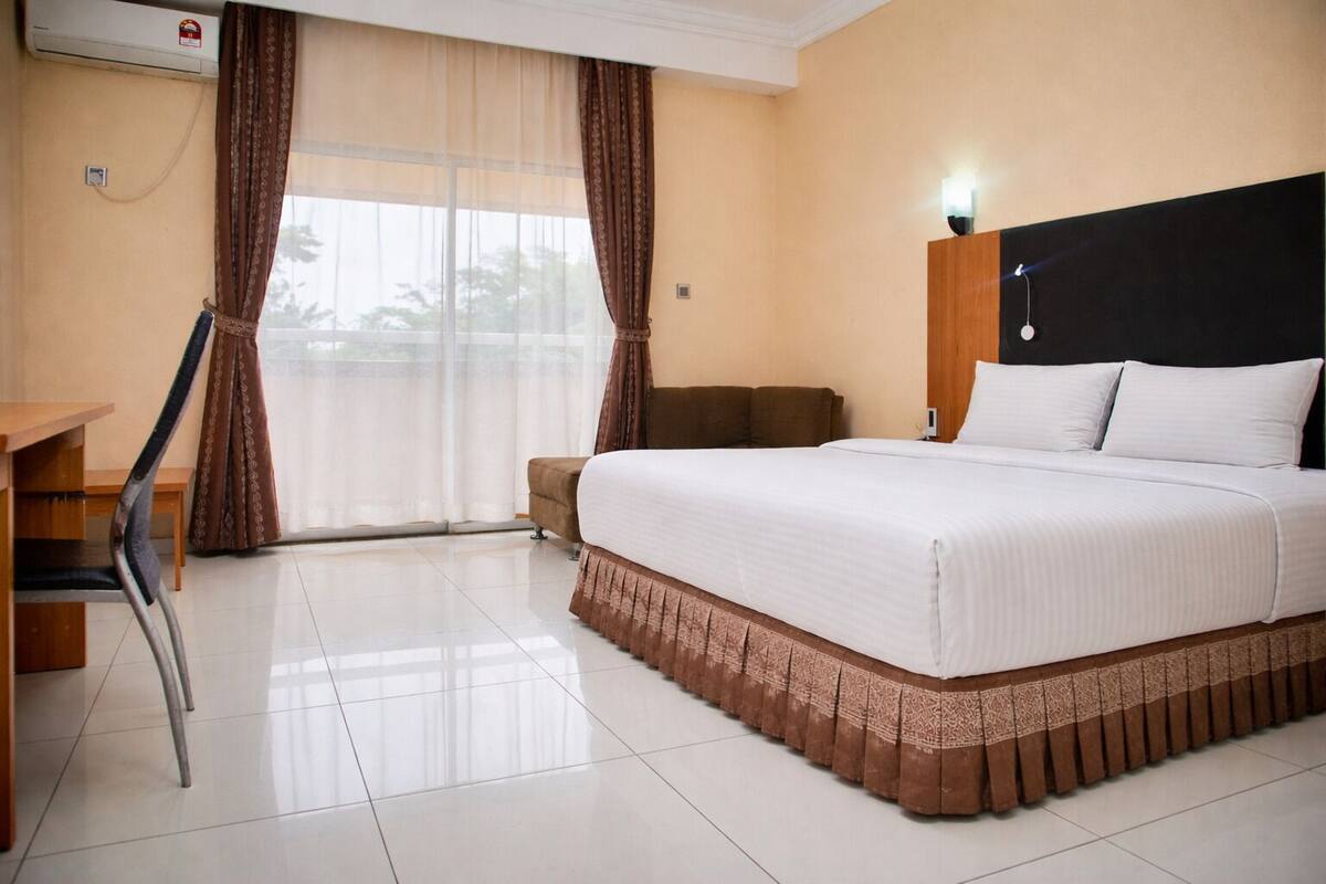 Quarto de hotel com uma cama, uma mesa, uma cadeira e uma janela com cortinas.