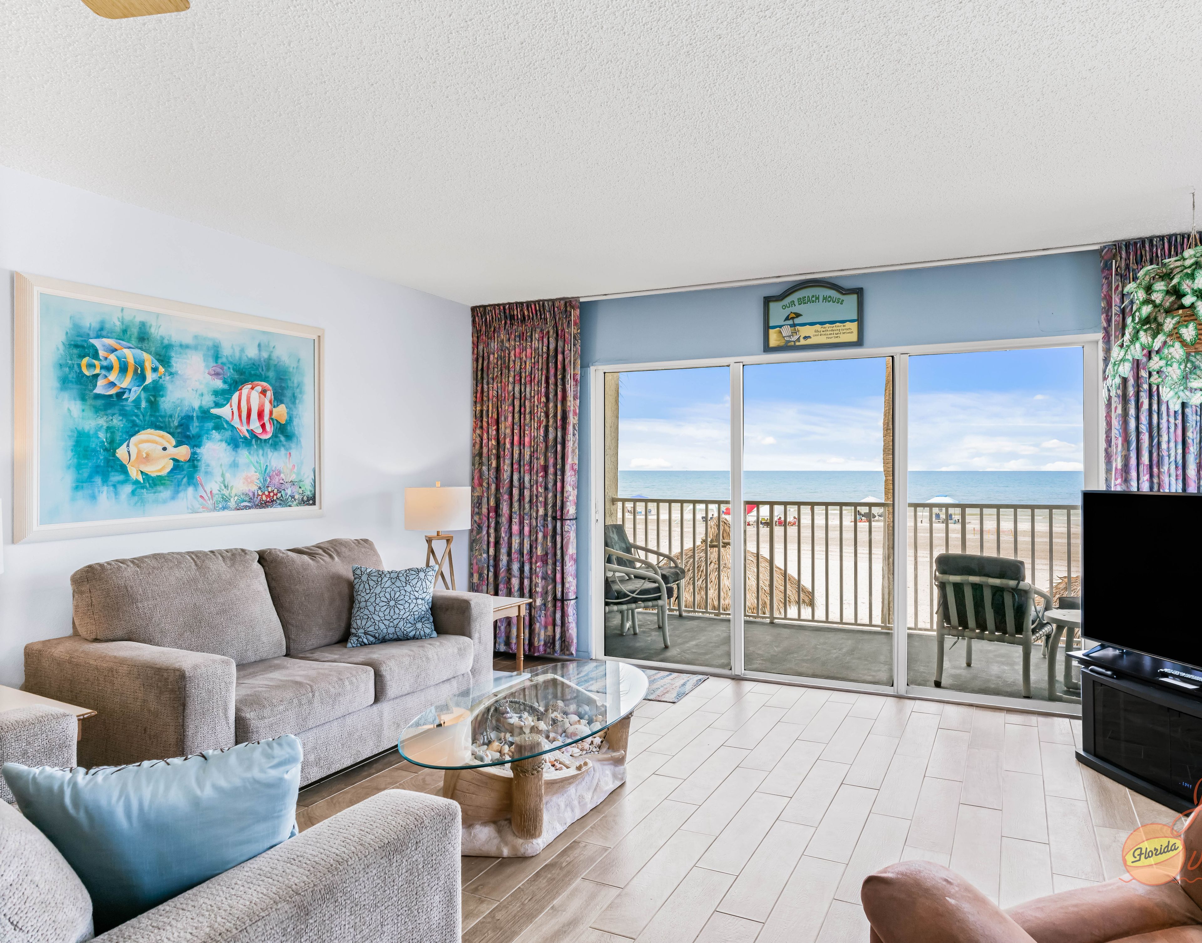 A living room with a sofa, armchairs, a coffee table, and a TV. There is a large window with a view of the beach.