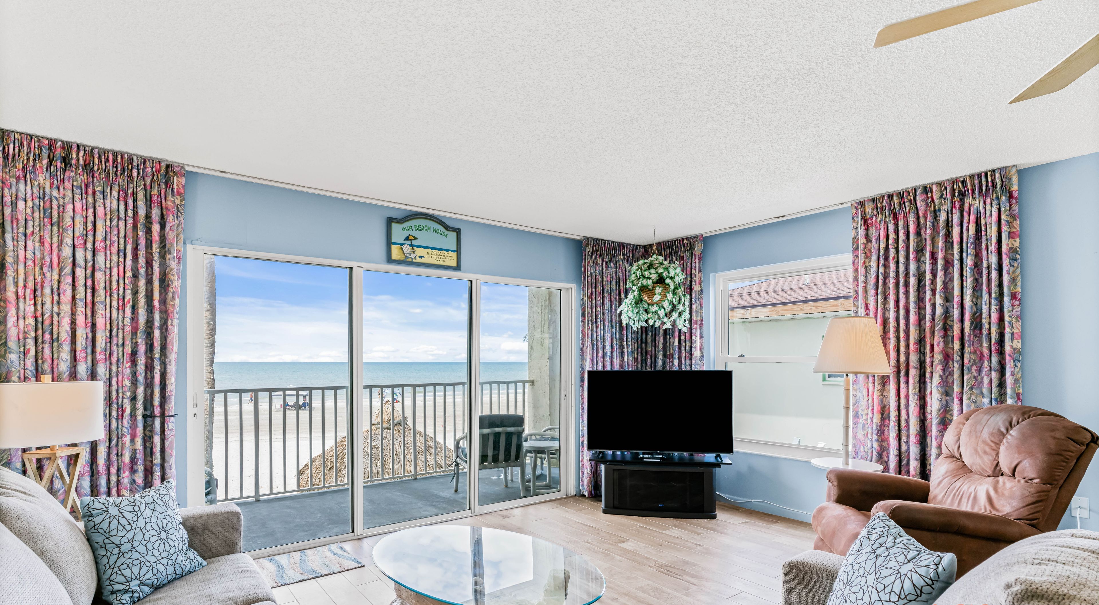 A living room with a glass coffee table, sofas, a TV, and a balcony with a view of the beach.