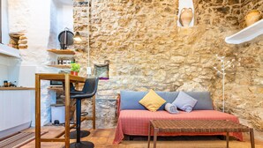 Una habitación acogedora con pared de piedra, cama con colcha roja y azul, una mesa de madera, una silla y una mesita con tapa de mimbre.