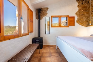 A bedroom with a bed, a bench, a window, a mirror, and a stone wall.