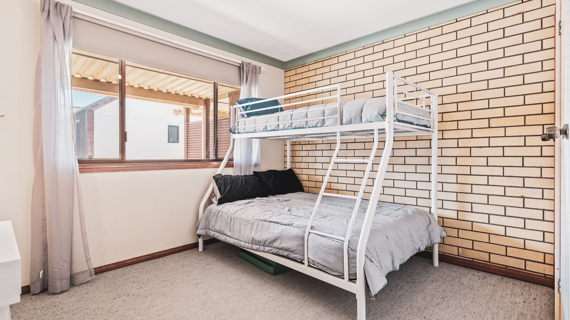 Een slaapkamer met een stapelbed, een bakstenen muur, een raam met gordijnen en uitzicht op een gebouw.