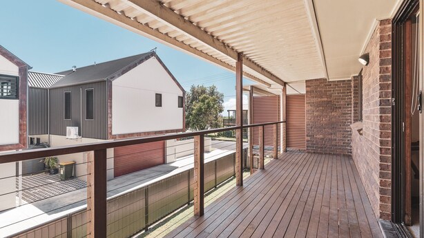 Un balcón techado con piso de madera, barandilla de metal y vista a casas vecinas.