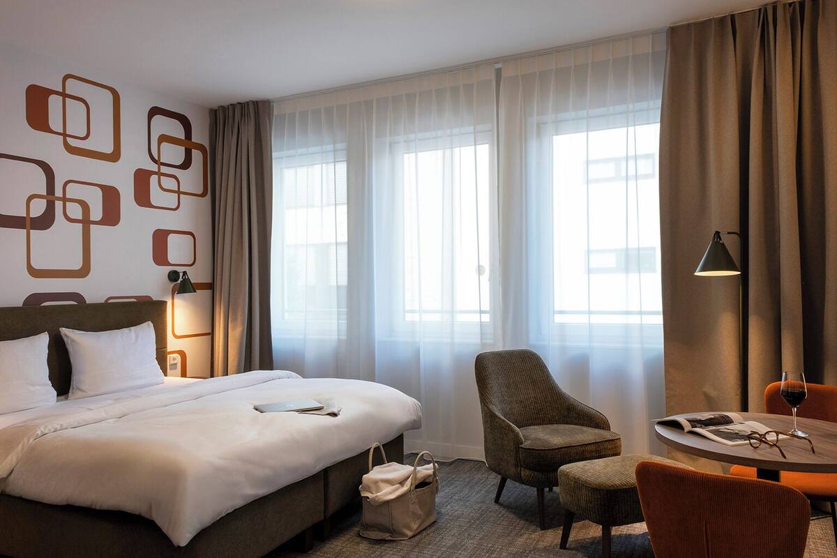 A hotel room with a bed, a chair, a small table, and a lamp.