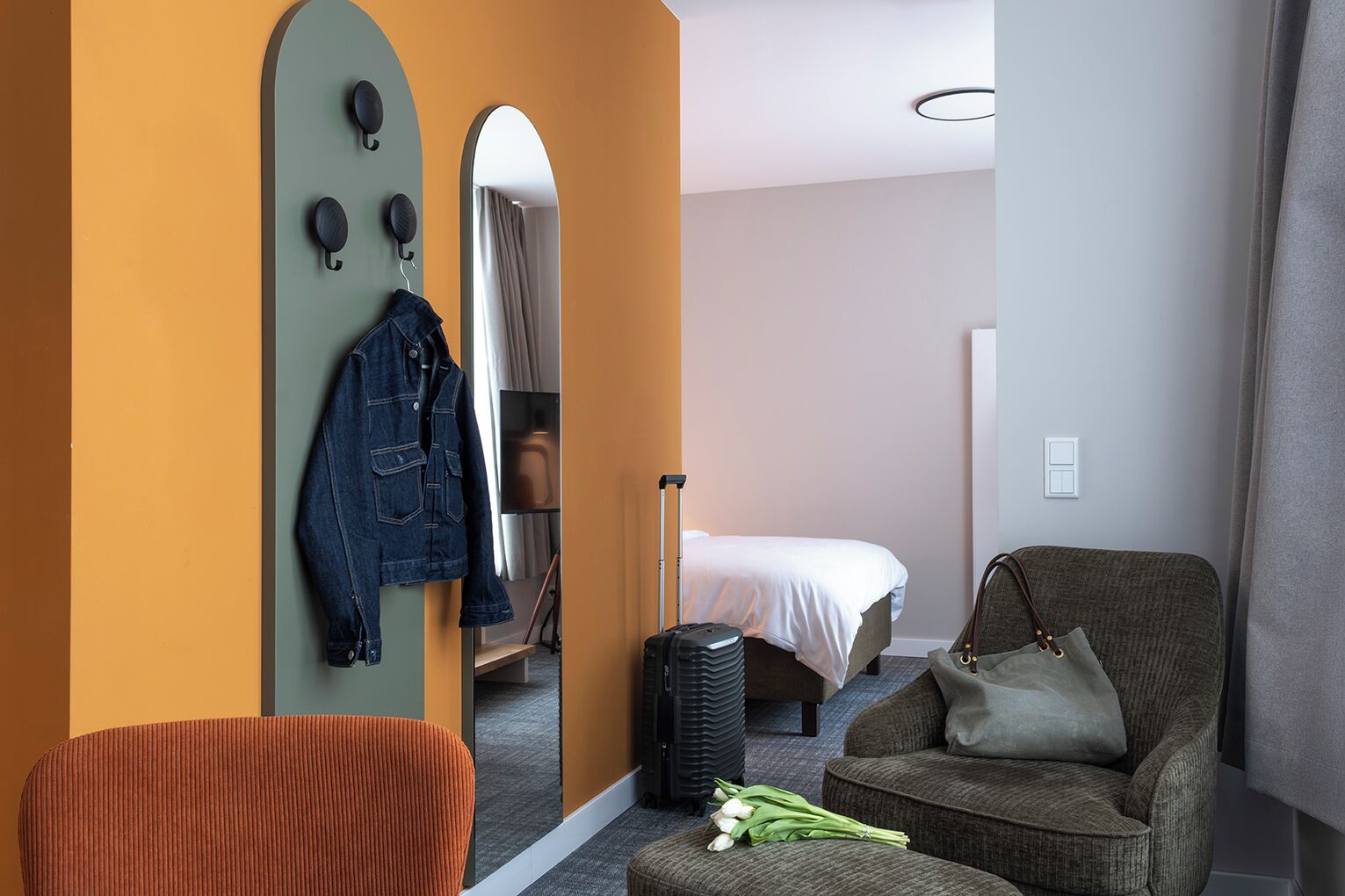 Una habitación de hotel con una cama, una zona de estar con un sillón, una maleta y una chaqueta colgada en la pared.