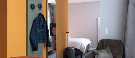 Una habitación de hotel con una cama, una zona de estar con un sillón, una maleta y una chaqueta colgada en la pared.