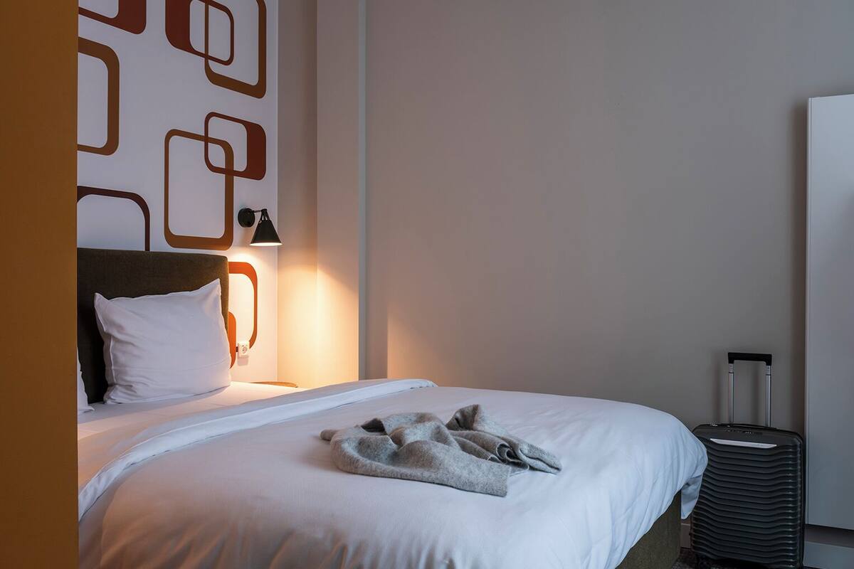 A hotel room with a neatly made bed, a wall-mounted lamp, and a suitcase.