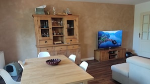 A living room with a wooden dining table, white chairs, a white sofa, a wooden cabinet with glass doors, and a television on a wooden stand.