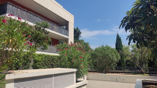 A modern building with balconies, a garden with flowering plants, and a clear blue sky.
