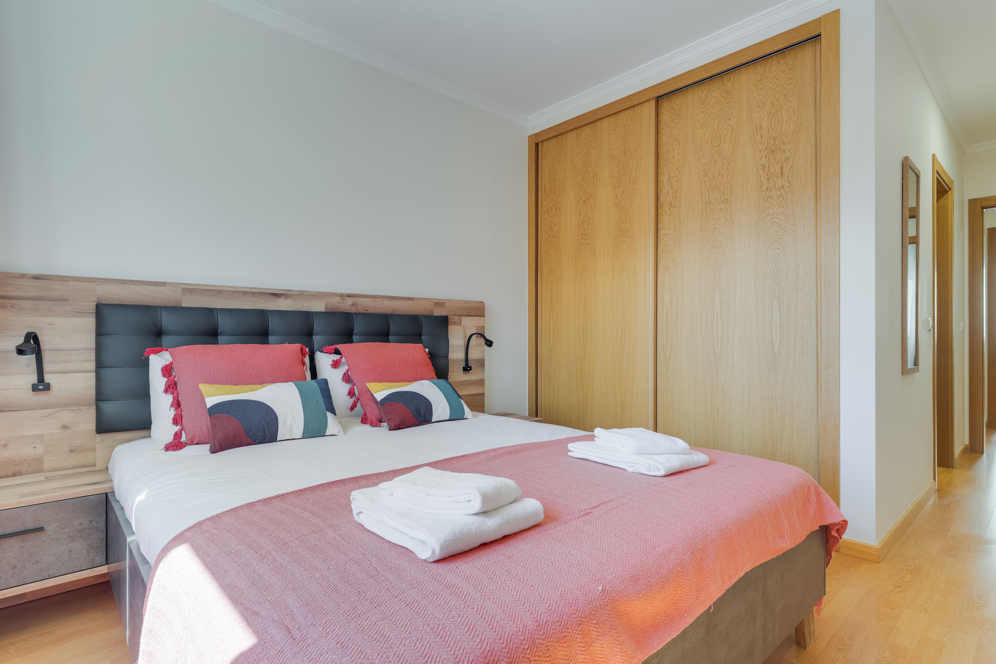A modern bedroom with a wooden headboard, a large bed with multiple pillows, a wooden wardrobe, and a hallway with a mirror.