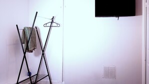 A room with a black TV mounted on the wall, a metal drying rack with clothes, and a vent on the wall.