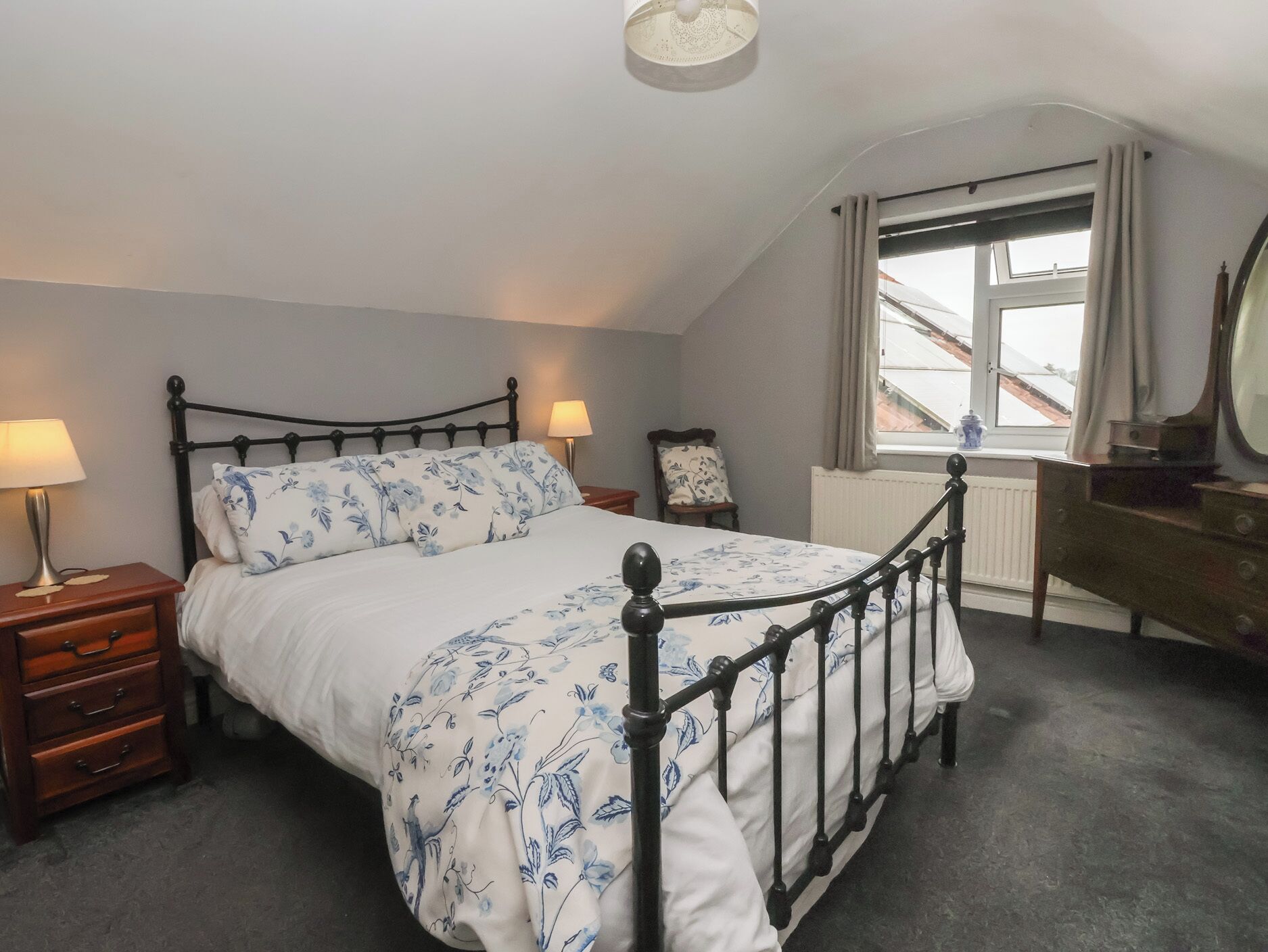 A bedroom with a bed, bedside tables, a chair, a desk, a mirror, and a window with a view.