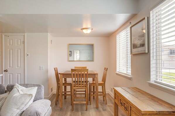 A dining area with a wooden table and chairs, a mirror on the wall, and a window with blinds.