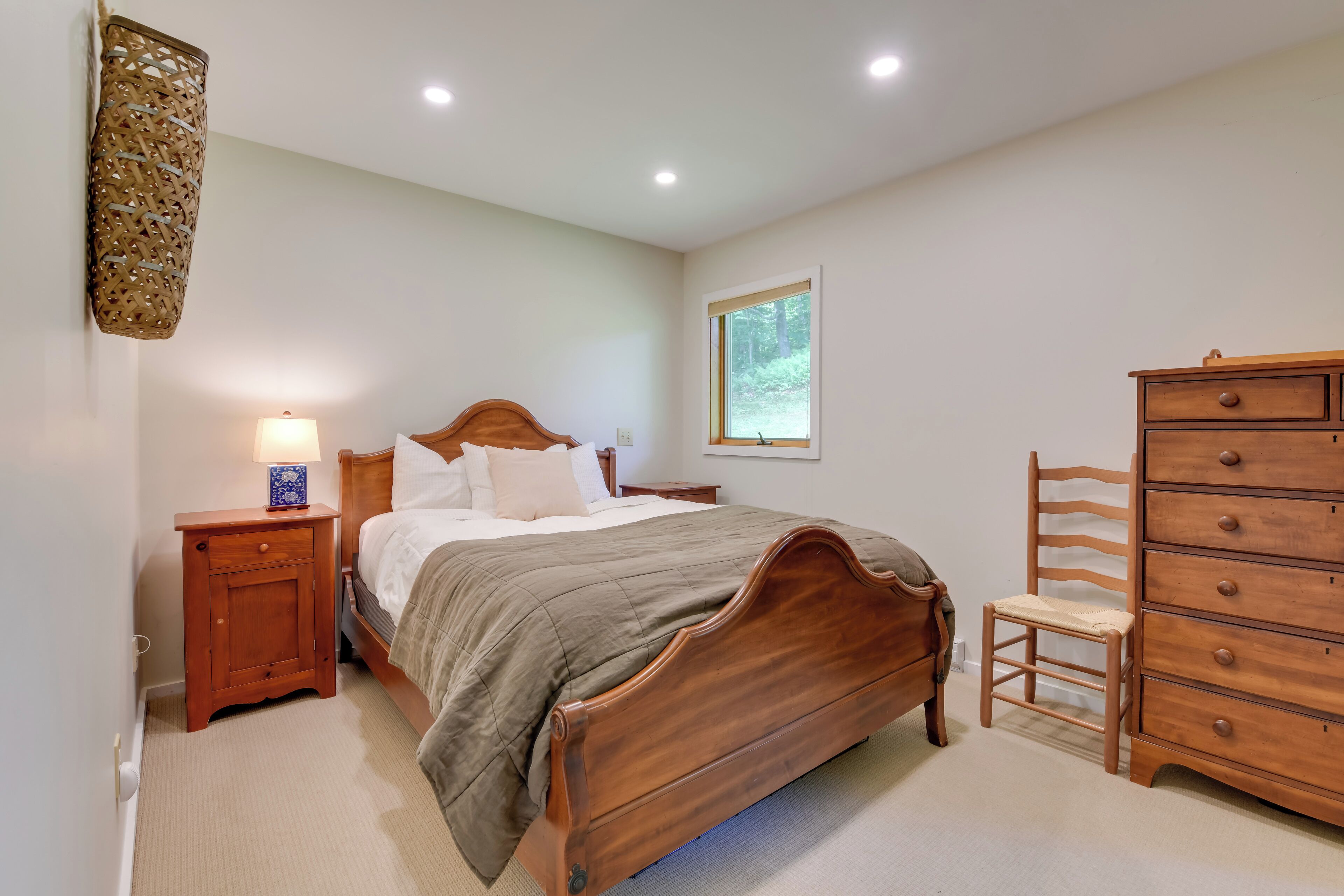 Une chambre à coucher avec un lit en bois, une table de chevet avec une lampe, une commode et une fenêtre donnant sur un paysage verdoyant.