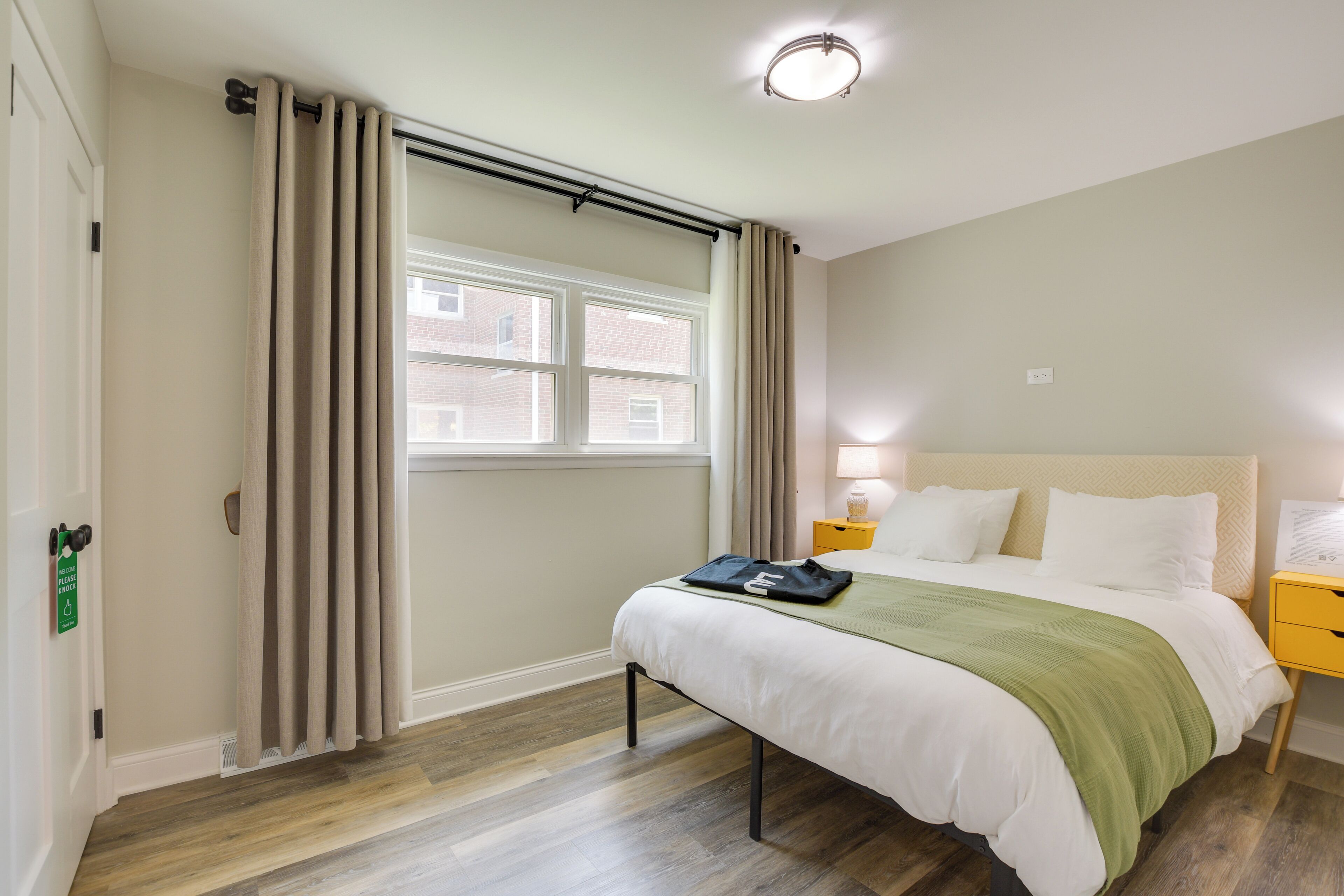 Una habitación de hotel con cama, mesita de noche, lámpara y ventana con cortinas.