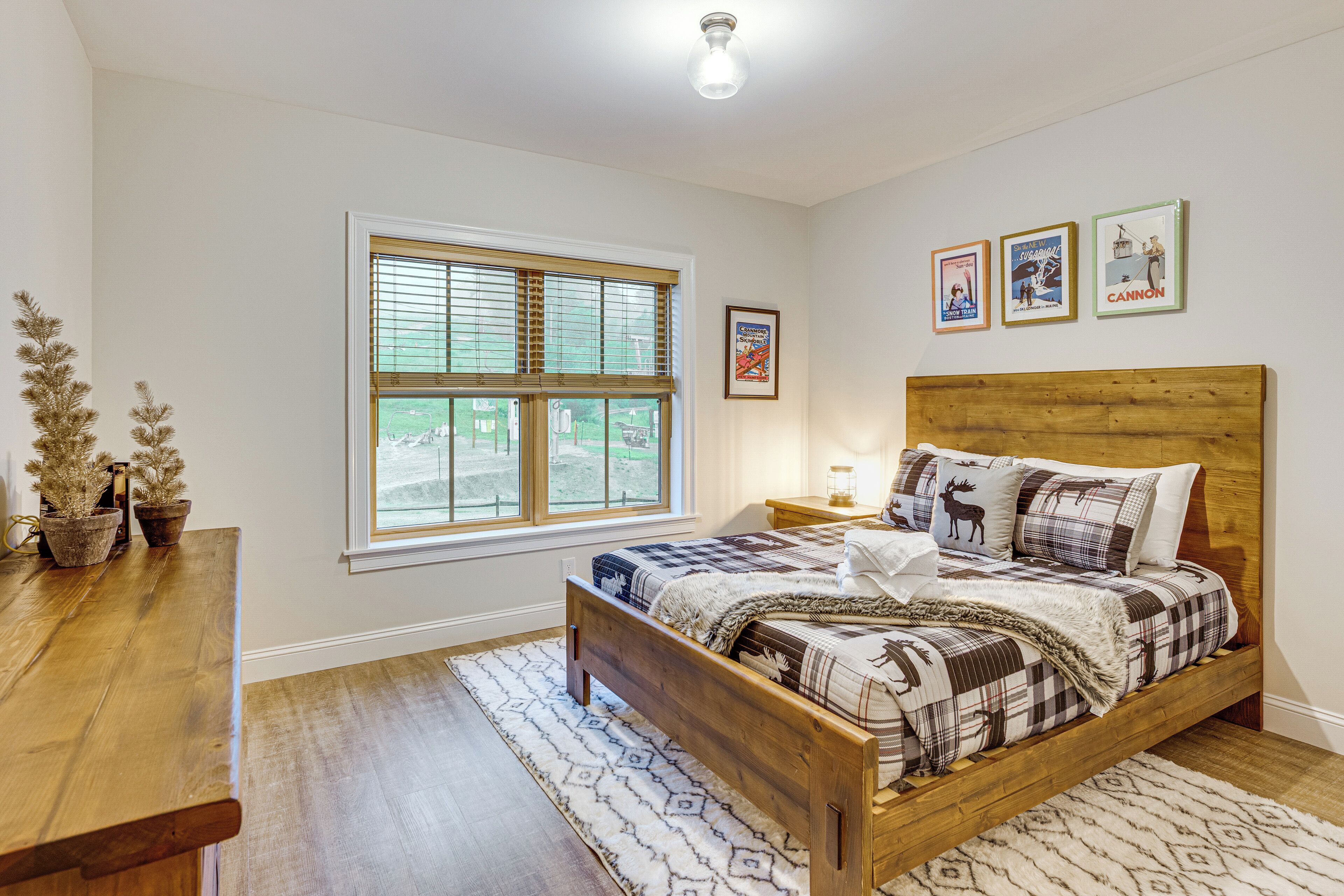A bedroom with a wooden bed, a window with blinds, a nightstand with a lamp, and a small table with potted plants.