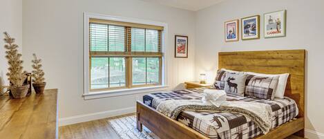 A bedroom with a wooden bed, a window with blinds, a nightstand with a lamp, and a small table with potted plants.