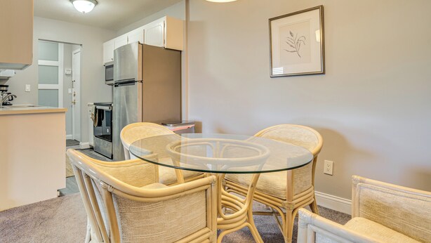 A dining area with a glass-top table surrounded by wicker chairs, a kitchen in the background, and a framed artwork on the wall. - Lake Access & Community Pool: Cozy Condo in Mound (Mound)