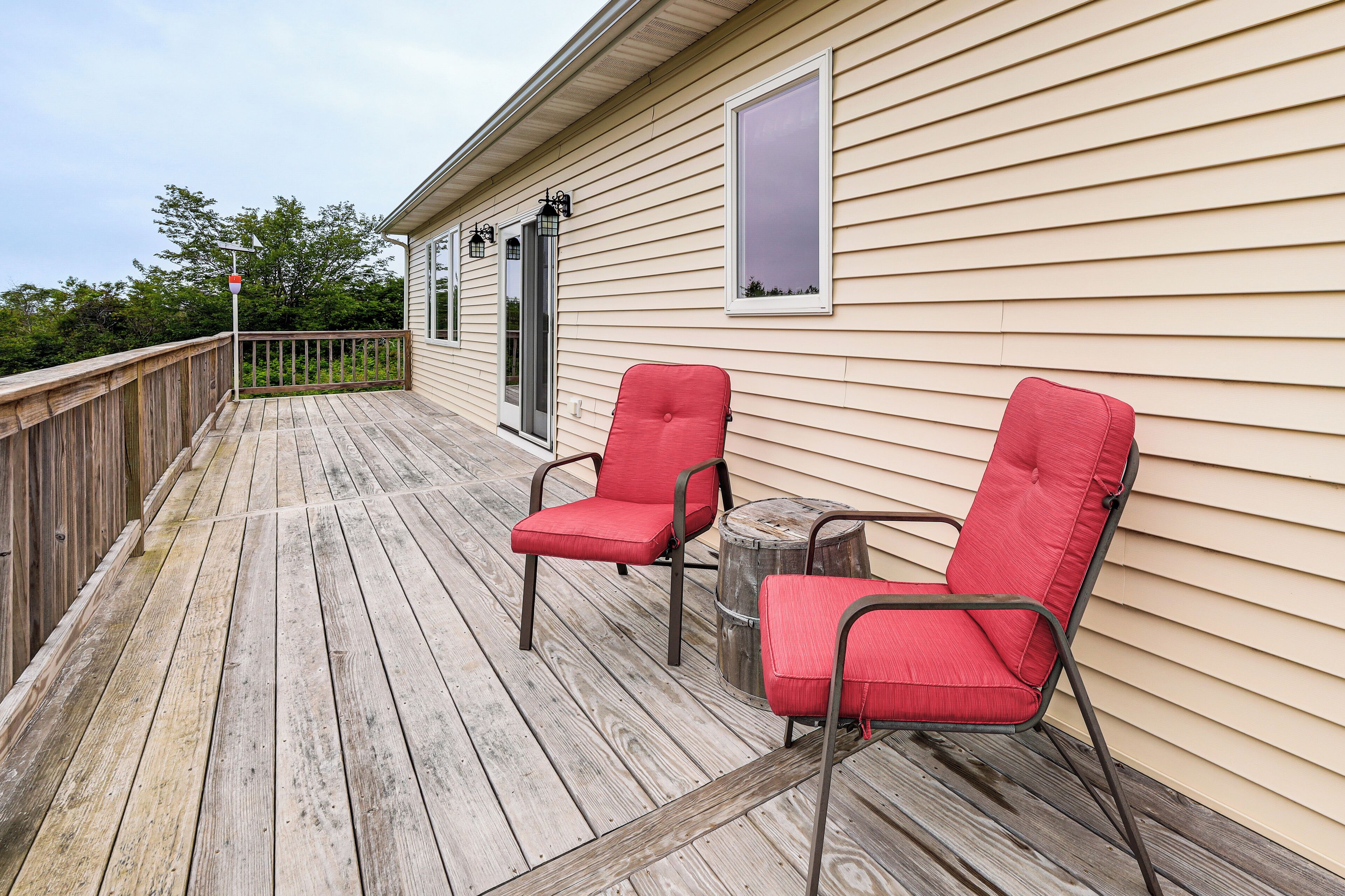 A wooden deck with two red chairs and a small table, a wooden railing, and a beige house with a window and a door.