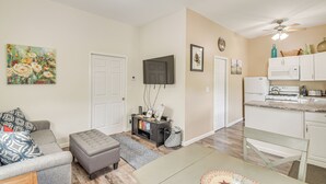 A living room with a grey sofa, a grey ottoman, and a TV on the wall. There is a kitchen area with white cabinets and a countertop.