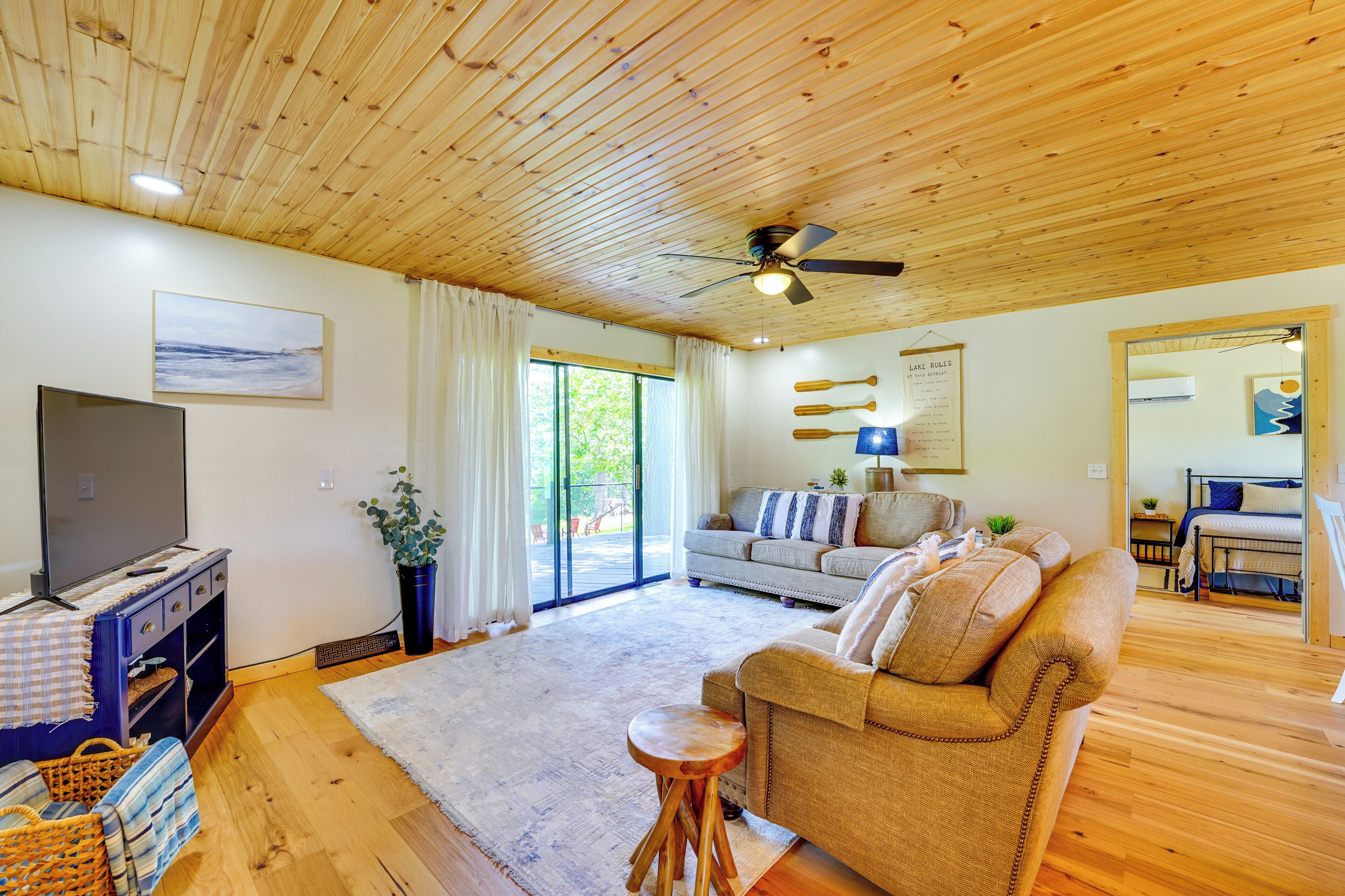 A cozy living room with a wooden ceiling, a flat-screen TV, a sofa, a coffee table, and a large window.