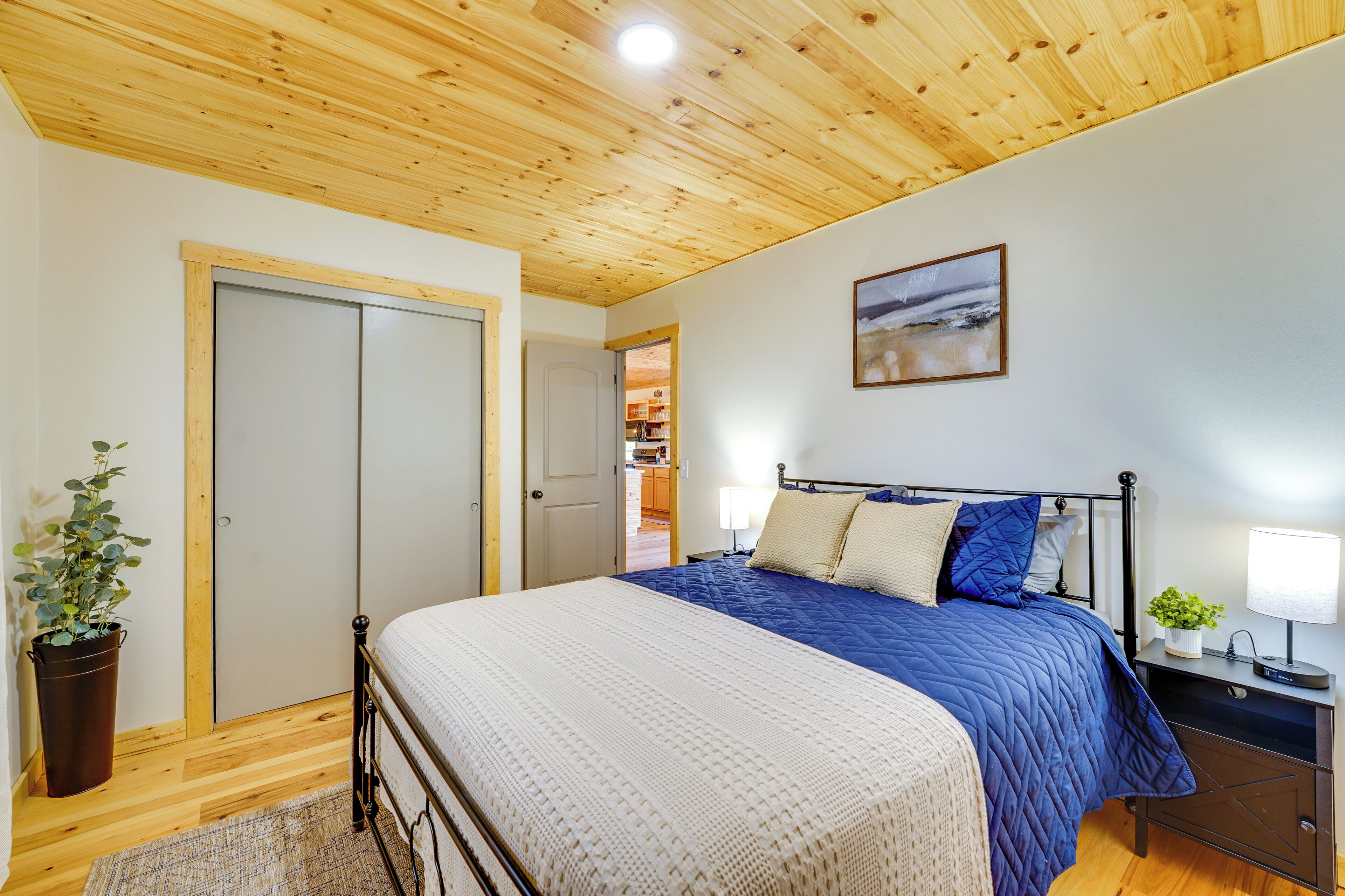 A bedroom with a wooden ceiling, a bed with blue and white bedding, a bedside table with a lamp, and a potted plant.