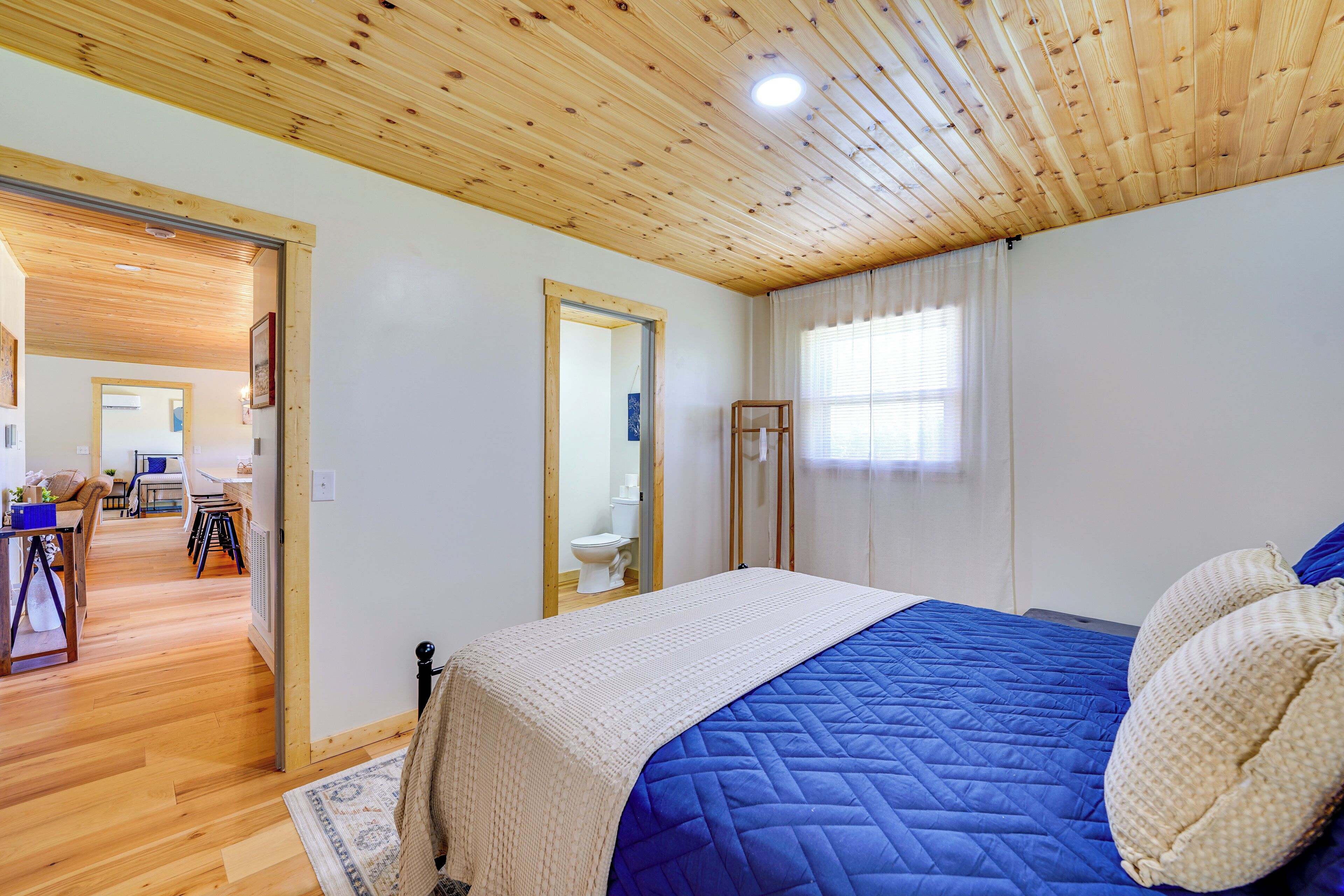 Un dormitorio con techo de madera, una cama acolchada azul, una manta blanca y una ventana con cortinas.