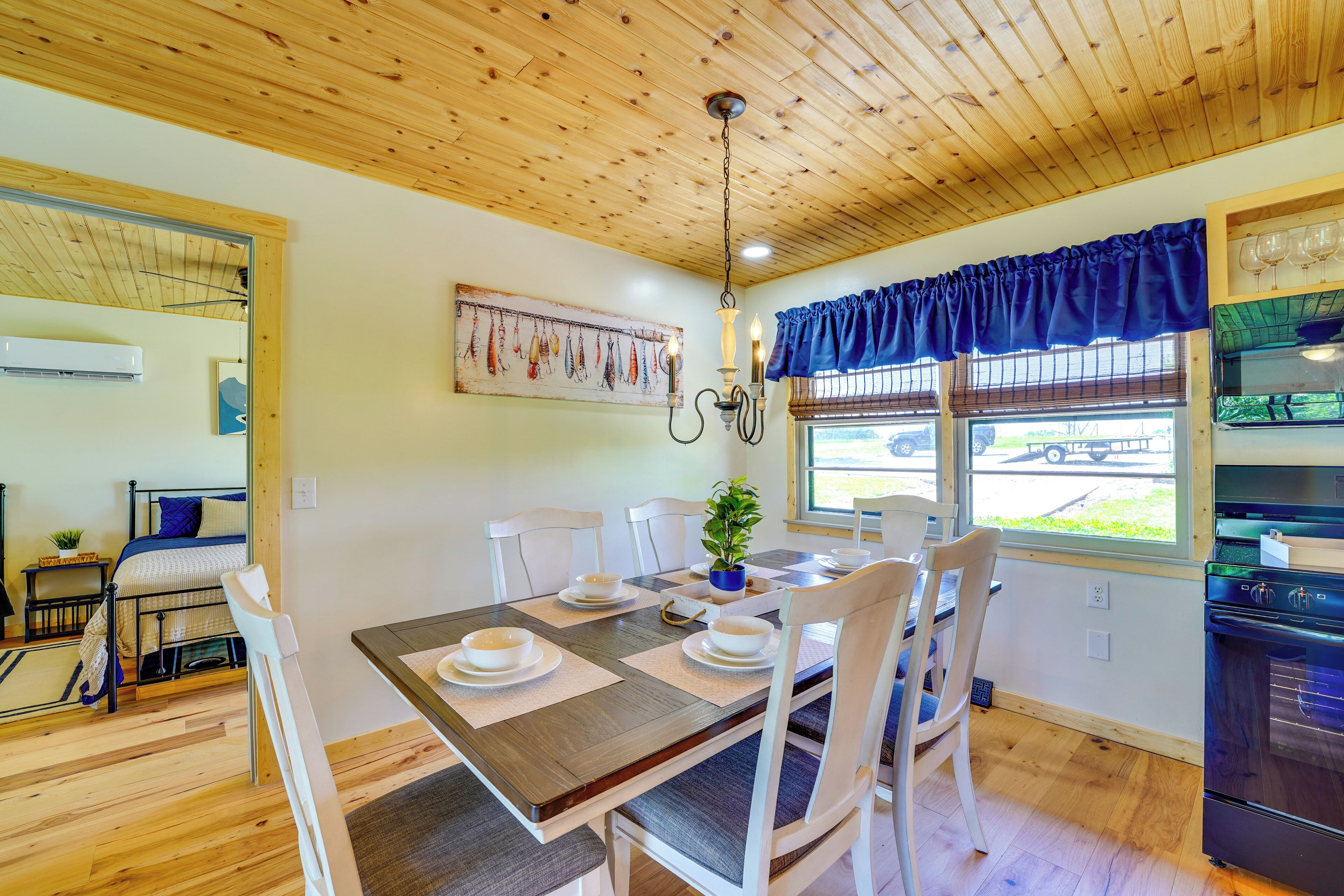 Un coin repas avec une table en bois dressée pour quatre personnes, entourée de chaises blanches, d’un lustre et d’une fenêtre ornée de rideaux bleus.