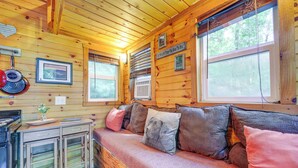 A wooden cabin interior with a wooden bench, a kitchen area with a stove and cabinets, and framed pictures on the walls. - Tiny Home w/ Private Sauna in Woods of Newport! (Newport)