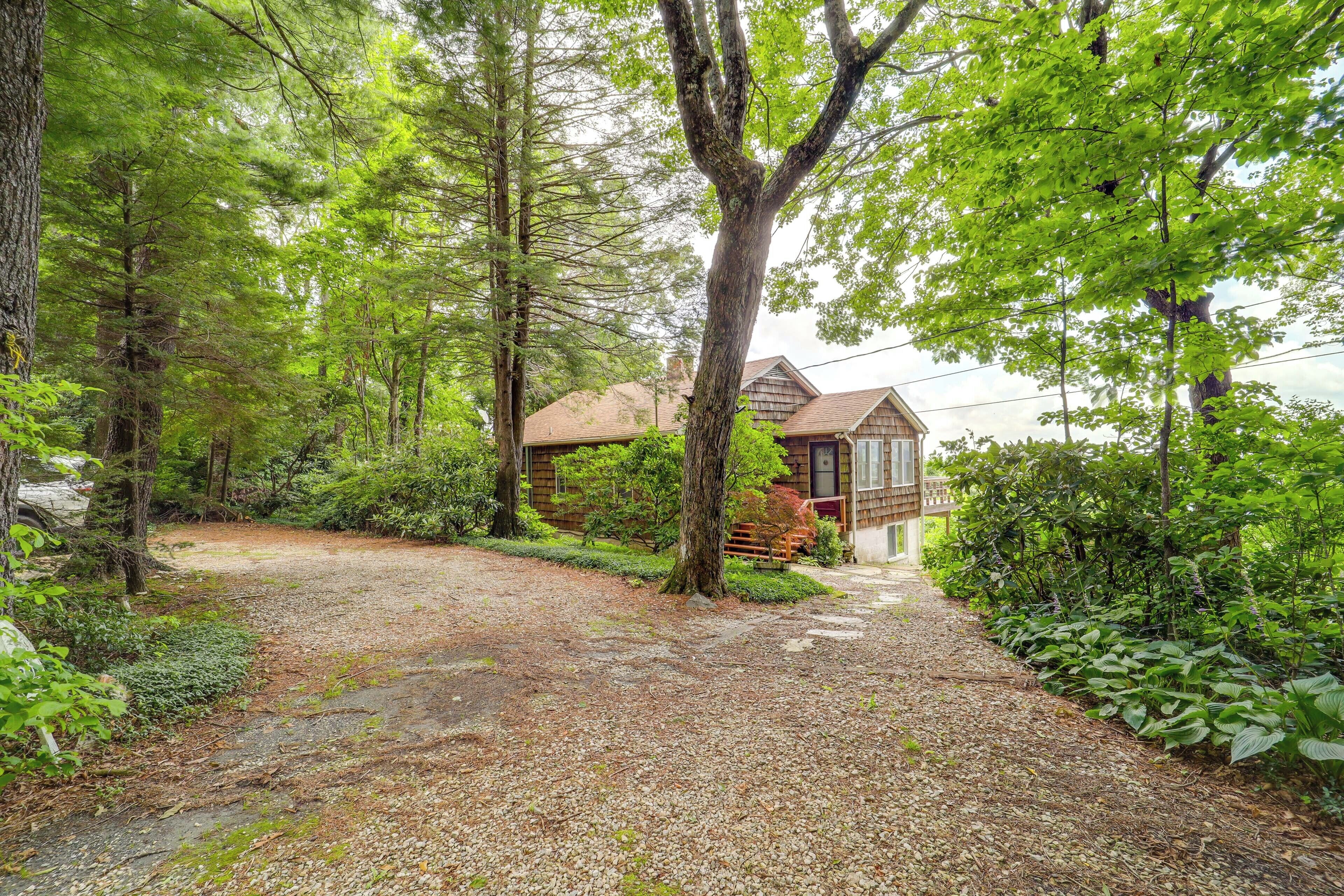 Un chalet rustique, entouré d’une forêt dense. Un sentier de gravier mène au chalet, qui est doté d’une terrasse en bois et d’un petit porche.