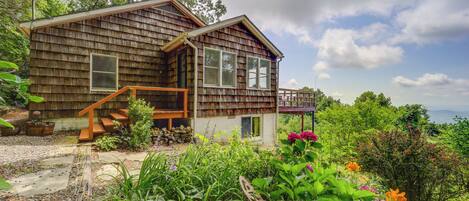 A wooden cabin with a porch, surrounded by lush greenery and flowers.