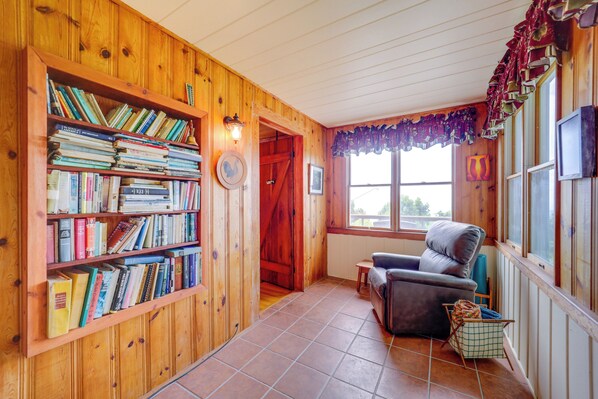 A cozy reading nook with a wooden bookshelf filled with books, a comfortable armchair, and a window with curtains. - Little Switzerland Cabin w/ Stunning Mtn Views (Little Switzerland)