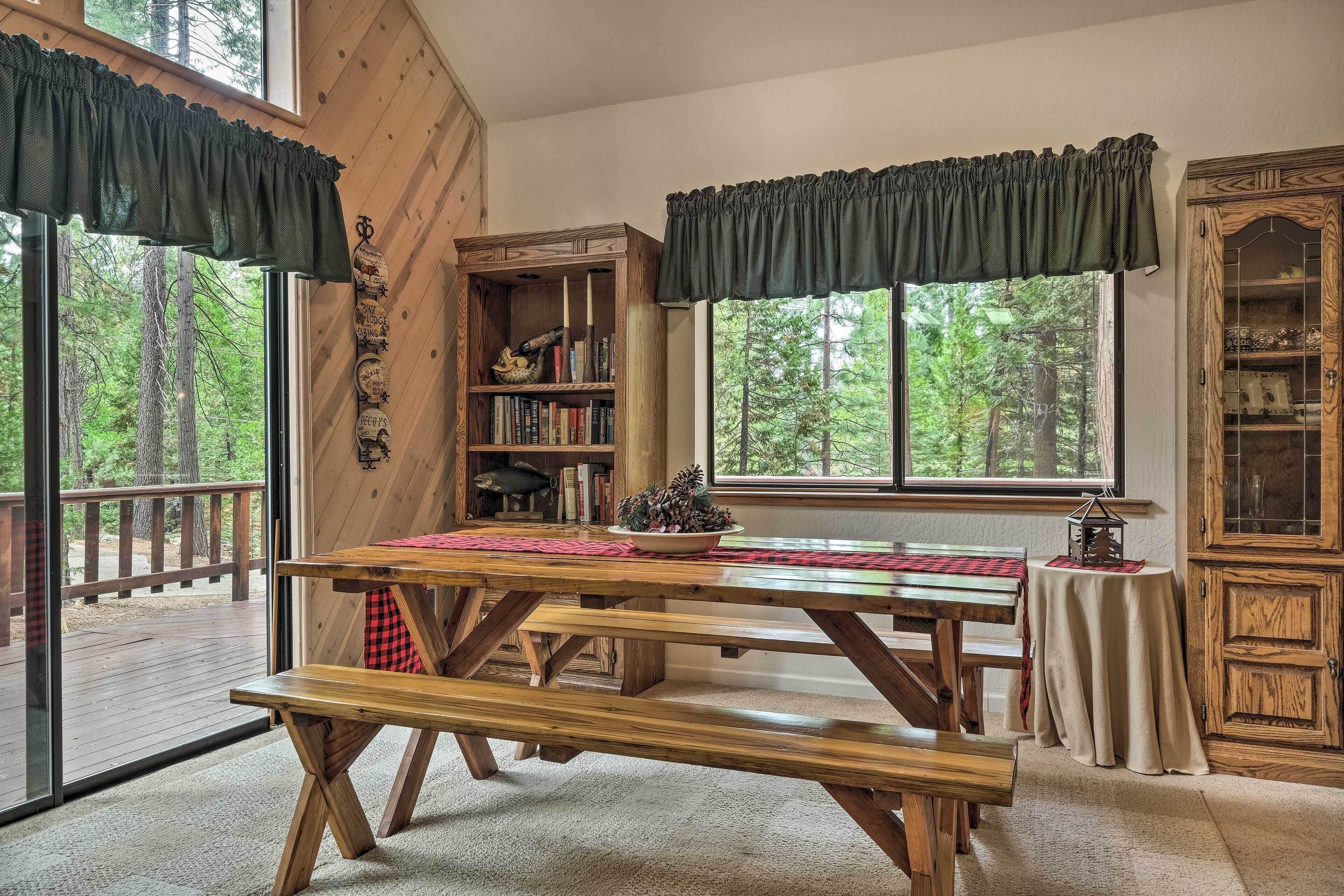 Een houten eettafel met banken, een boekenkast en een kast in een kamer met grote ramen en uitzicht op bomen.