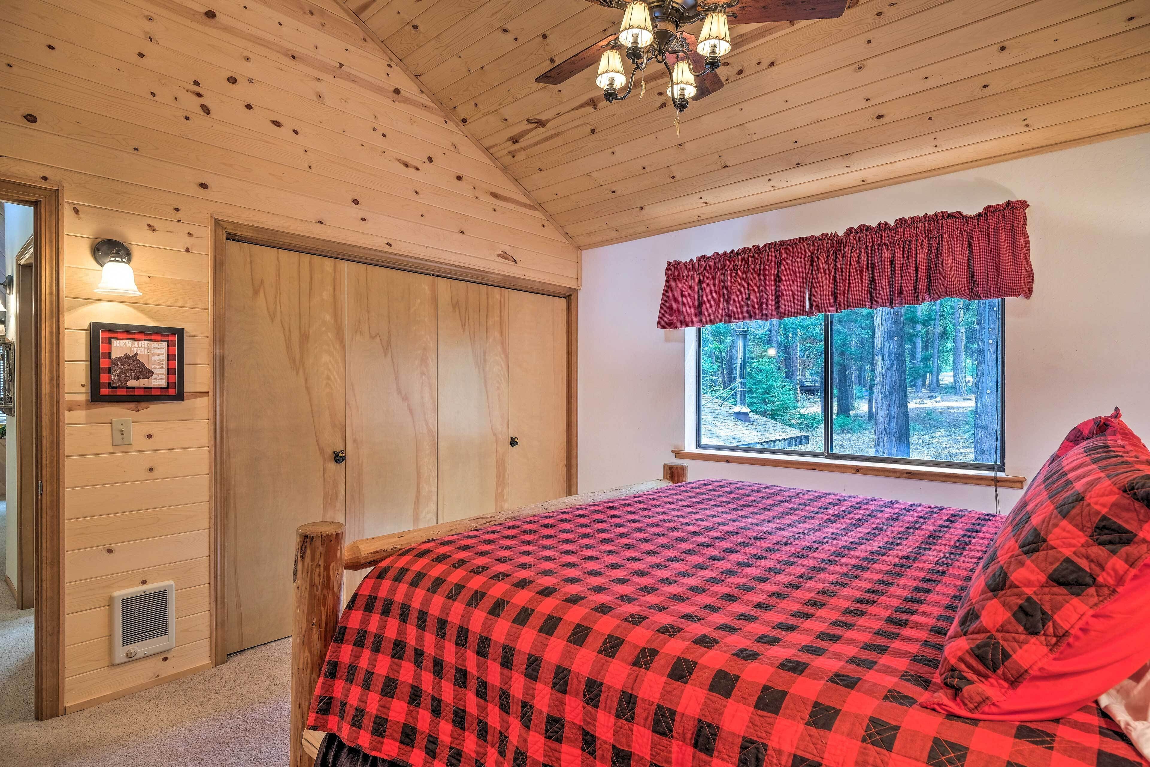 Un dormitorio con techo de madera, una cama con colcha de cuadros rojos y negros, una ventana con cortina roja y una puerta con interruptor de luz y rejilla de ventilación.
