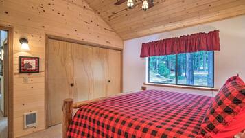 Un dormitorio con techo de madera, una cama con colcha de cuadros rojos y negros, una ventana con cortina roja y una puerta con interruptor de luz y rejilla de ventilación.