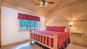 A bedroom with a wooden ceiling, a bed with a red and black checkered bedspread, a window with a red valance, and two wall-mounted lamps. - Game Room & Deck: Pet-friendly Camp Connell Cabin (Arnold)