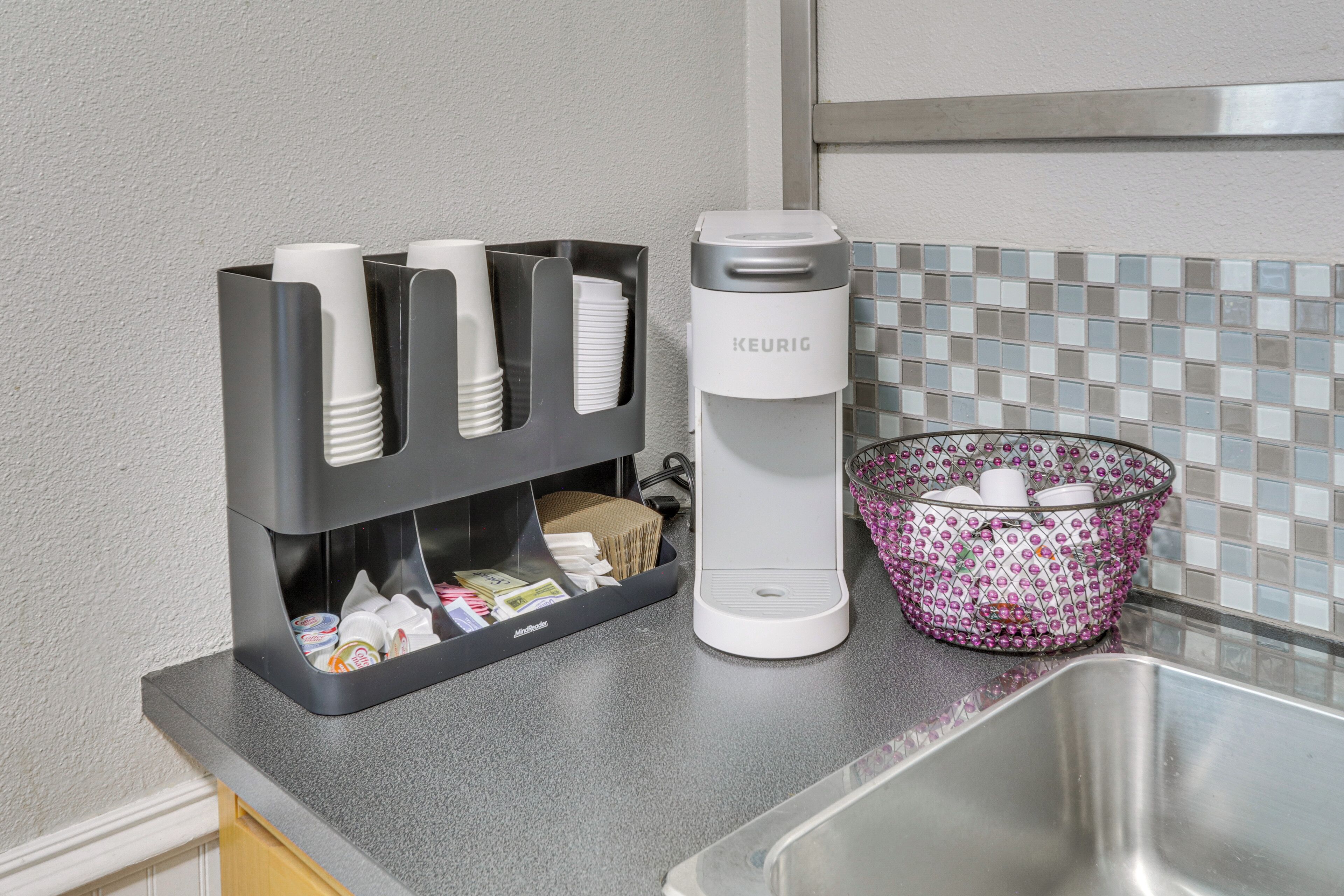 A kitchen counter with a coffee maker, a cup holder with disposable cups, and a basket with cups.
