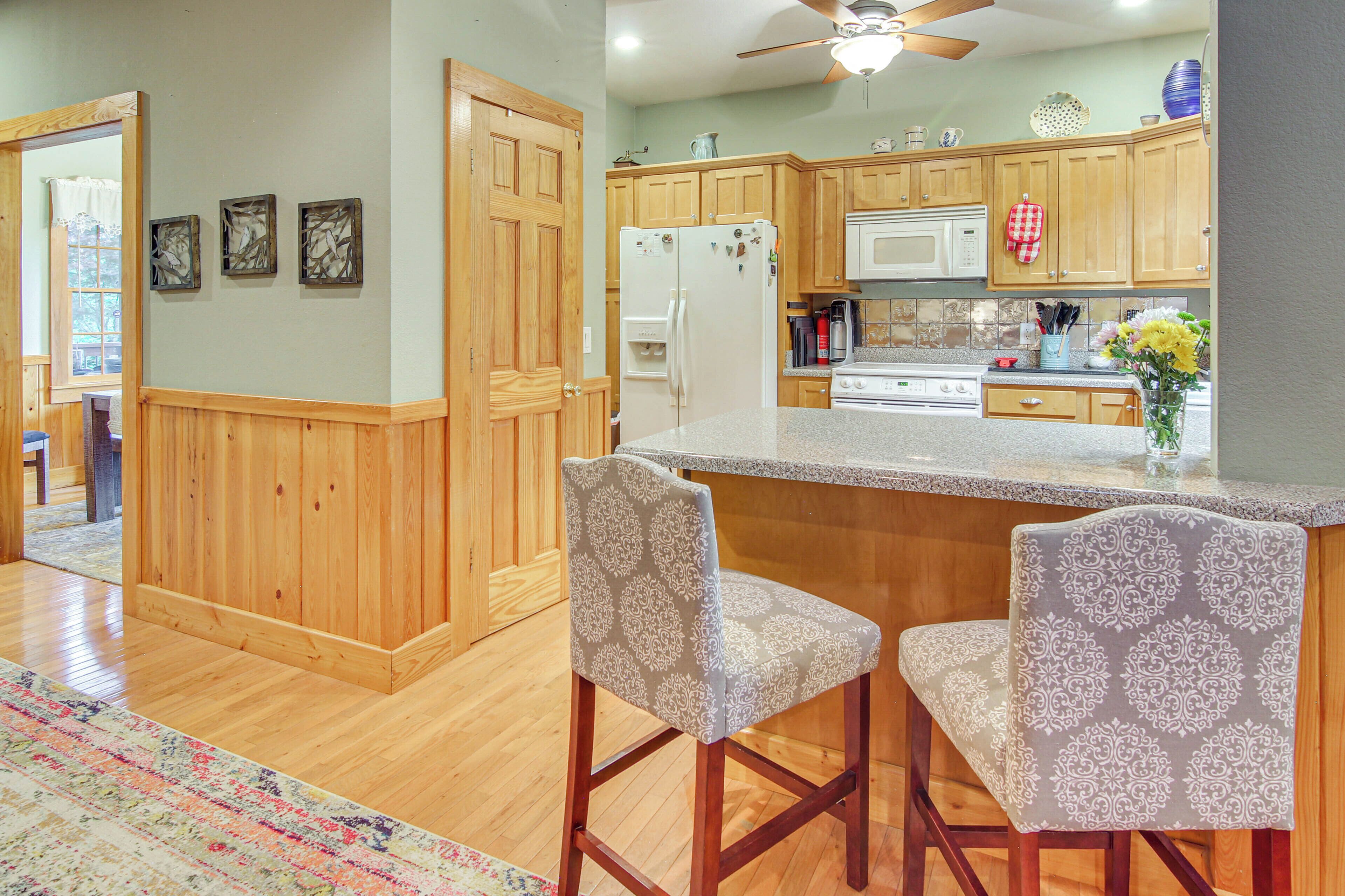 A kitchen with wooden cabinets, a refrigerator, a microwave, and a stove. There are two bar stools with patterned upholstery.