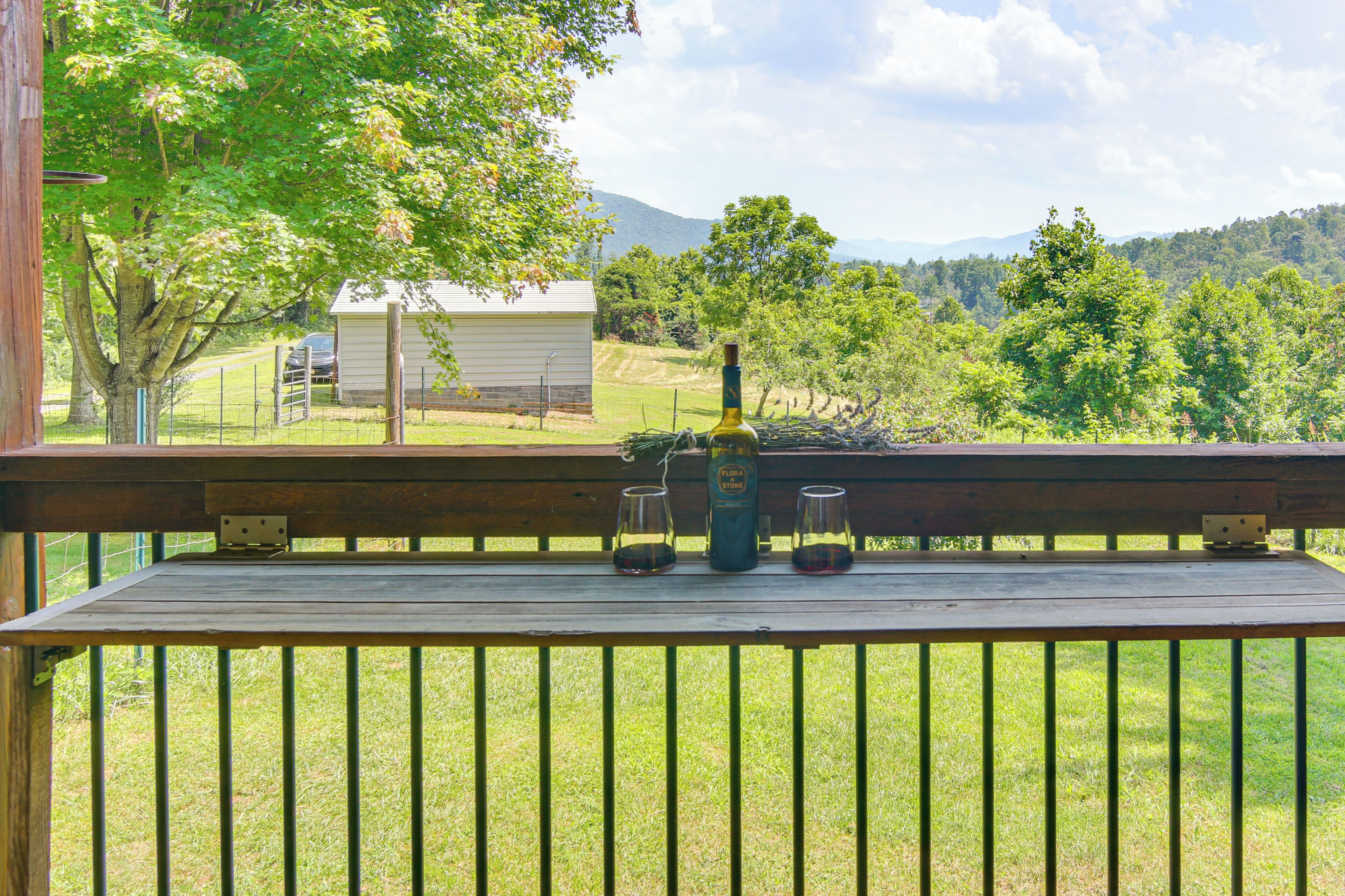 A wooden deck with a bottle of wine and two glasses on a table, overlooking a grassy area with trees and a distant building.