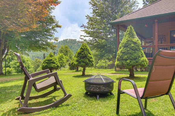 Une terrasse avec un fauteuil à bascule, un foyer et une chaise offrant une vue sur un chalet et les arbres.