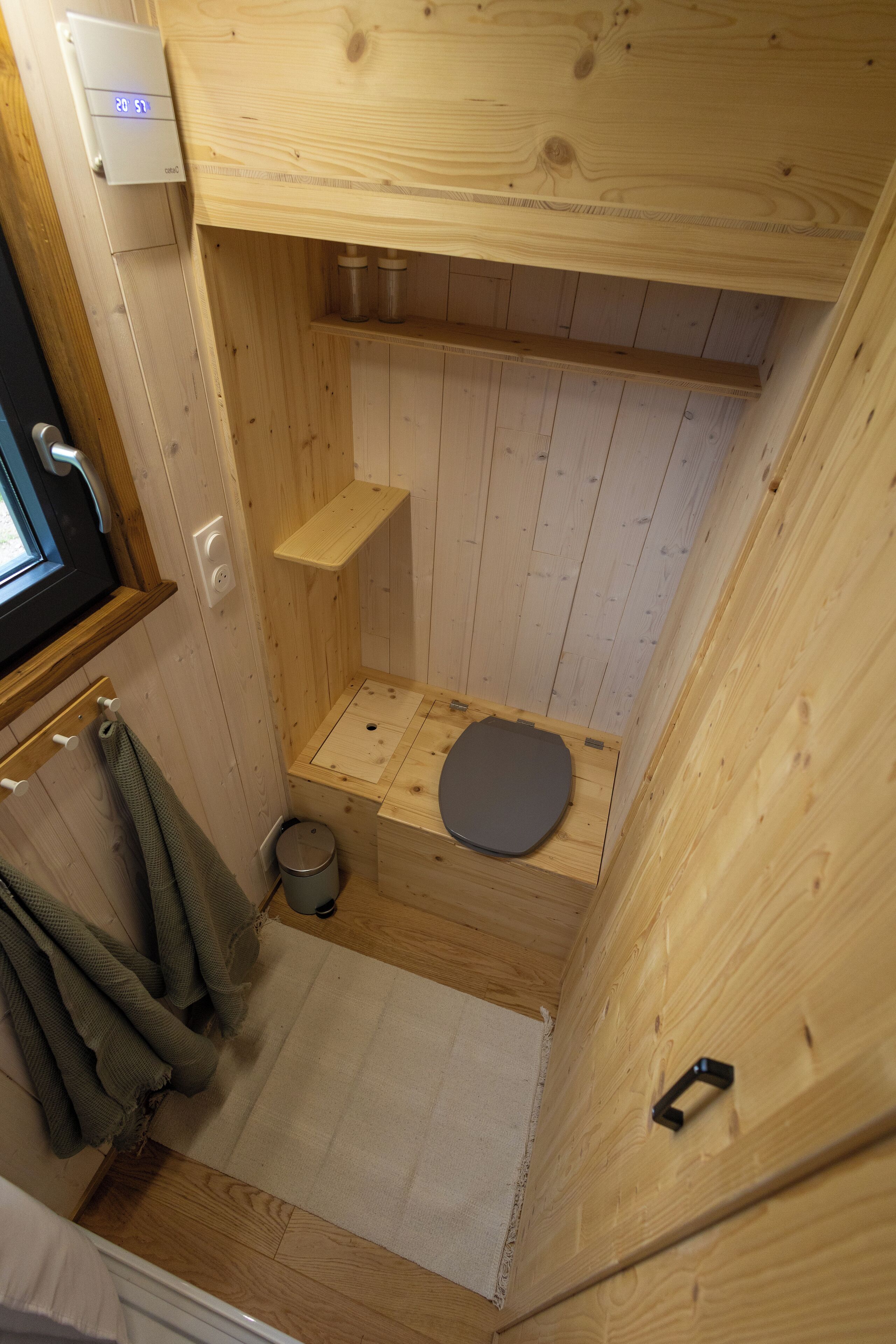 Una pequeña habitación de madera con un inodoro, un estante, una toalla y una ventana.
