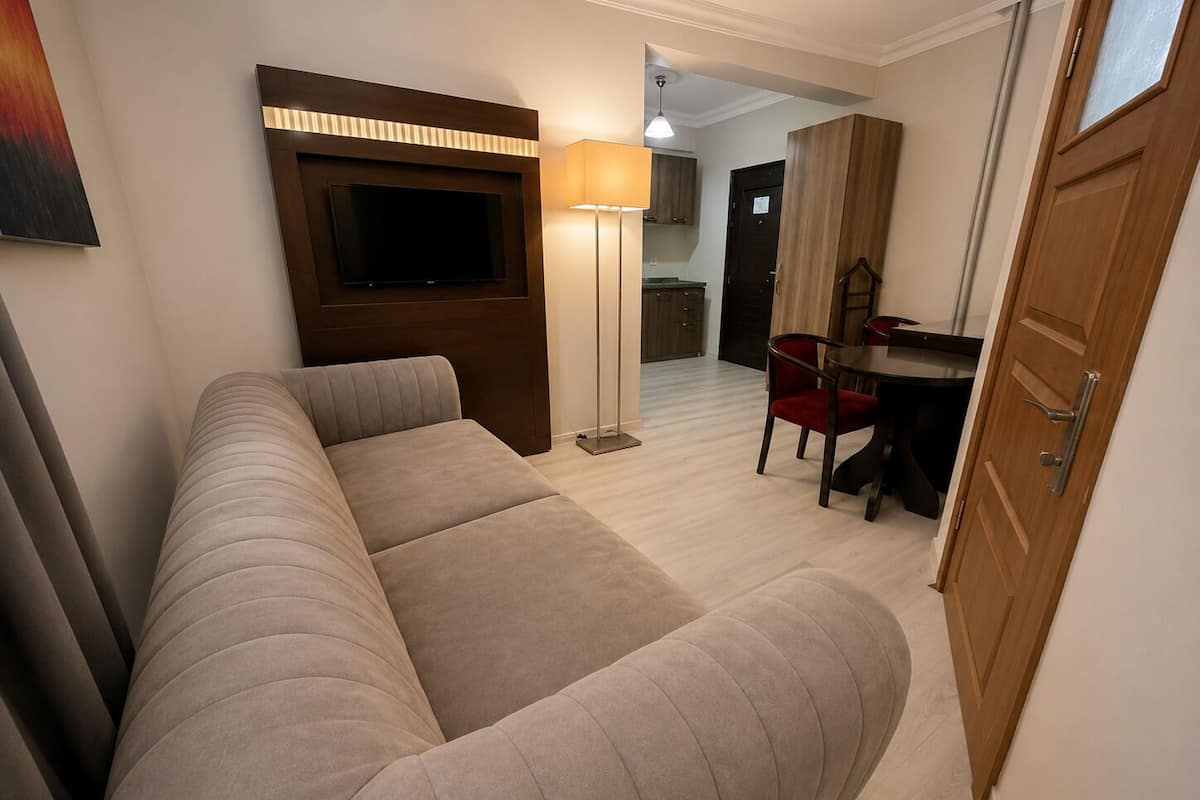 A modern hotel room with a sofa, a TV mounted on the wall, a dining area with a table and chairs, and a wooden door.