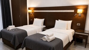 A hotel room with two beds, each with a dark grey bedspread, white pillows, and rolled towels. There are wall-mounted lamps on either side of the beds, and a small bedside table with a lamp on the right.