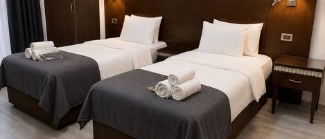 Habitación de hotel con dos camas, cada una con colcha gris oscuro, almohadas blancas y toallas enrolladas. Hay lámparas de pared a cada lado de las camas y una mesita de noche pequeña con una lámpara a la derecha.