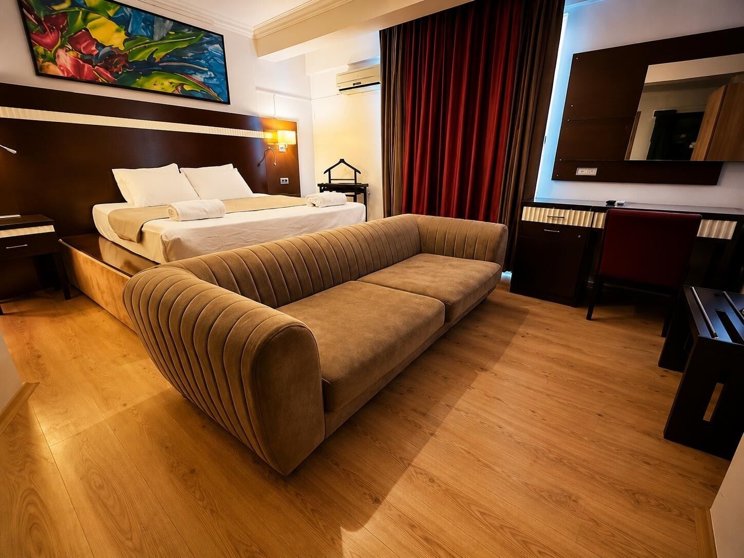 A hotel room with a bed, a sofa, a desk, a chair, and a painting on the wall.