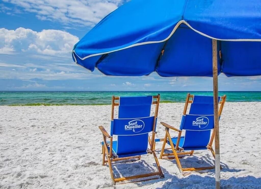 Zwei blaue Strandstühle mit 'Surf Dueller'-Branding unter einem blauen Sonnenschirm auf einem Sandstrand, im Hintergrund ein klarer Himmel und ein Meer.