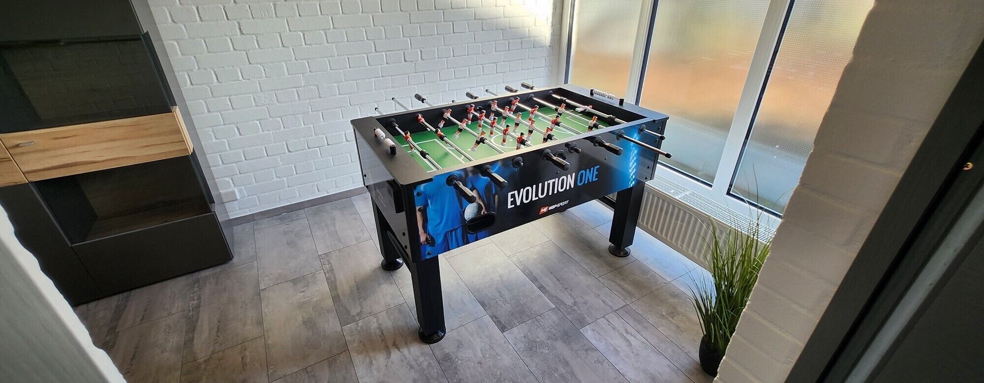 A foosball table labeled 'EVOLUTION ONE' in a room with a white brick wall, a wooden cabinet, and a view through a window.