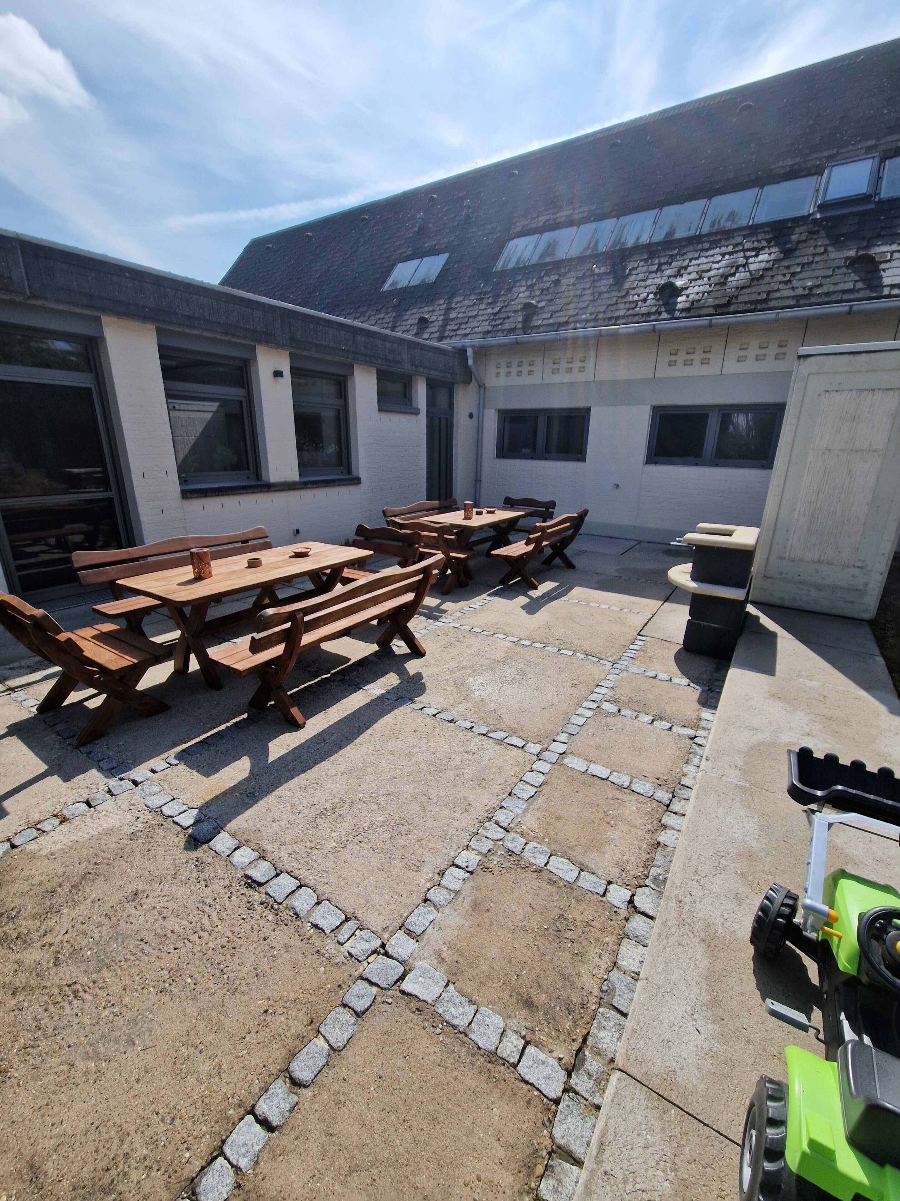 Un patio al aire libre con mesas y bancos de madera, suelo de concreto con bordes de adoquines, y un cortacésped verde en primer plano.