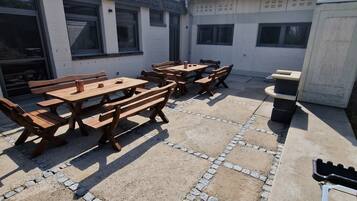 Un patio al aire libre con mesas y bancos de madera, suelo de concreto con bordes de adoquines, y un cortacésped verde en primer plano.